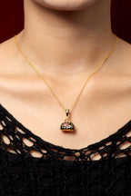 A gold chain necklace with a small handbag-shaped pendant featuring black enamel and a clear rhinestone center, worn by a model in a black lace top against a dark red background.