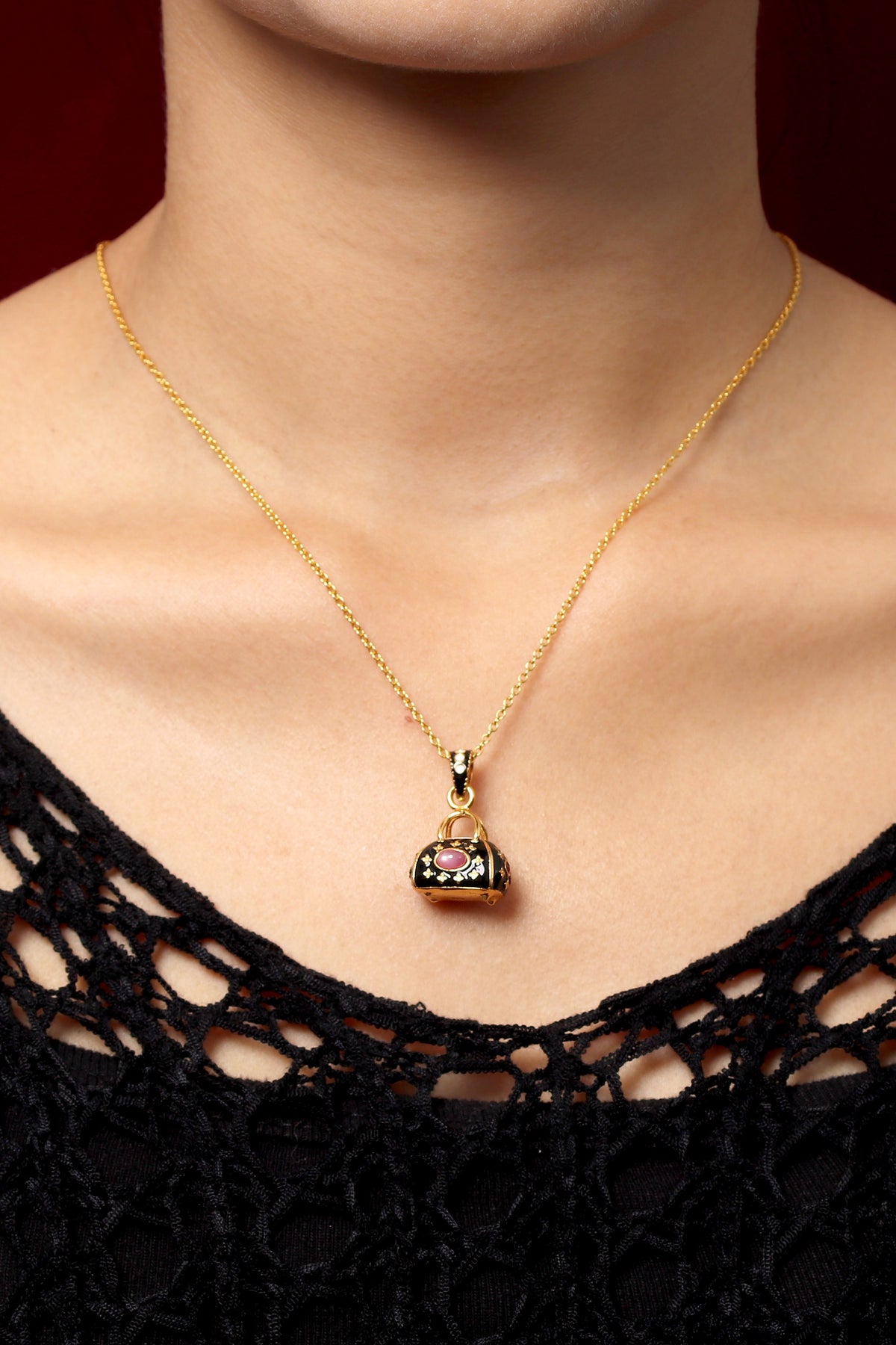 A gold chain necklace with a small handbag-shaped pendant featuring black enamel and a clear rhinestone center, worn by a model in a black lace top against a dark red background.