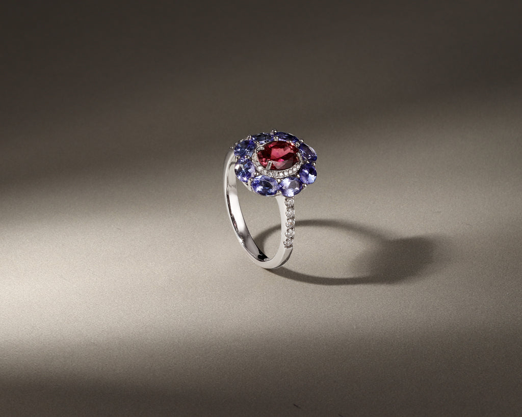 A silver ring featuring a central round red gemstone surrounded by a halo of blue gemstones and small clear accent stones, displayed on a neutral gray background with dramatic lighting.