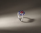 A silver ring featuring a central round red gemstone surrounded by a halo of blue gemstones and small clear accent stones, displayed on a neutral gray background with dramatic lighting.