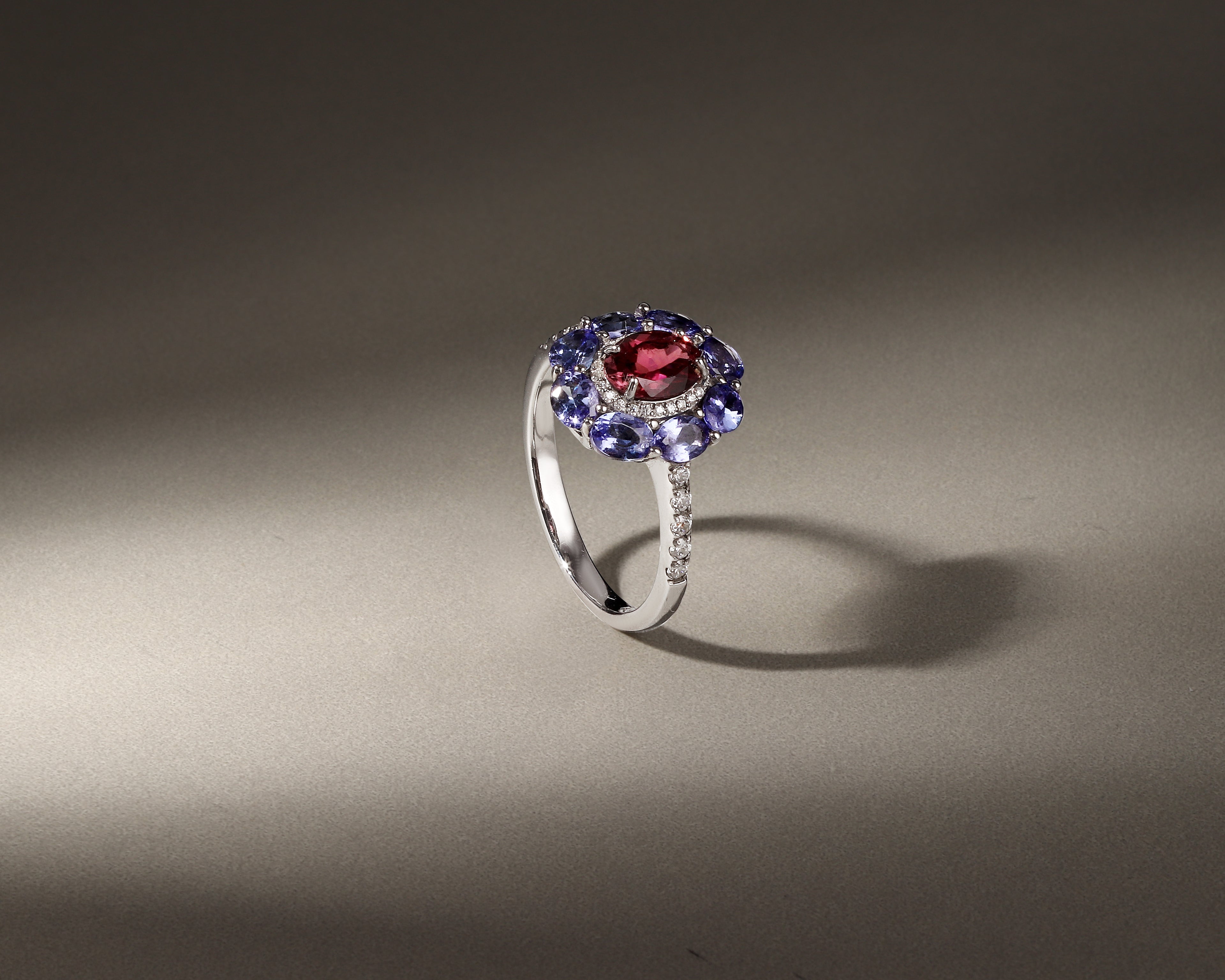 A silver ring featuring a central round red gemstone surrounded by a halo of blue gemstones and small clear accent stones, displayed on a neutral gray background with dramatic lighting.