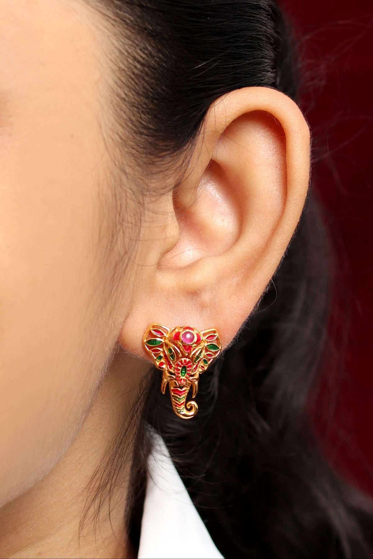 A close-up of a person's ear wearing a small elephant-shaped stud earring with gold-tone metal and red and green enamel details, against a dark red background.