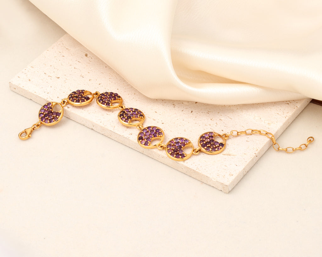 A gold-tone bracelet with five oval links set with sparkling purple crystals, displayed on a textured cream stone slab with draped satin fabric.