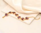 A gold-tone bracelet with five oval links set with sparkling purple crystals, displayed on a textured cream stone slab with draped satin fabric.