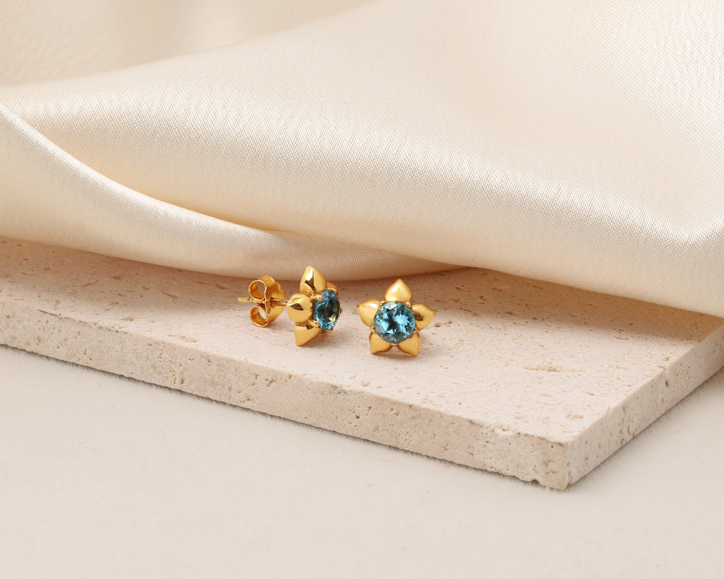 A pair of gold flower-shaped stud earrings with round blue gemstones, displayed on a textured beige stone slab with draped cream satin fabric in the background.