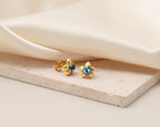 A pair of gold flower-shaped stud earrings with round blue gemstones, displayed on a textured beige stone slab with draped cream satin fabric in the background.