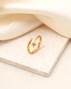 A gold ring with a twisted open band and a small green star-shaped gemstone, displayed on a textured beige stone slab with draped cream fabric in the background.