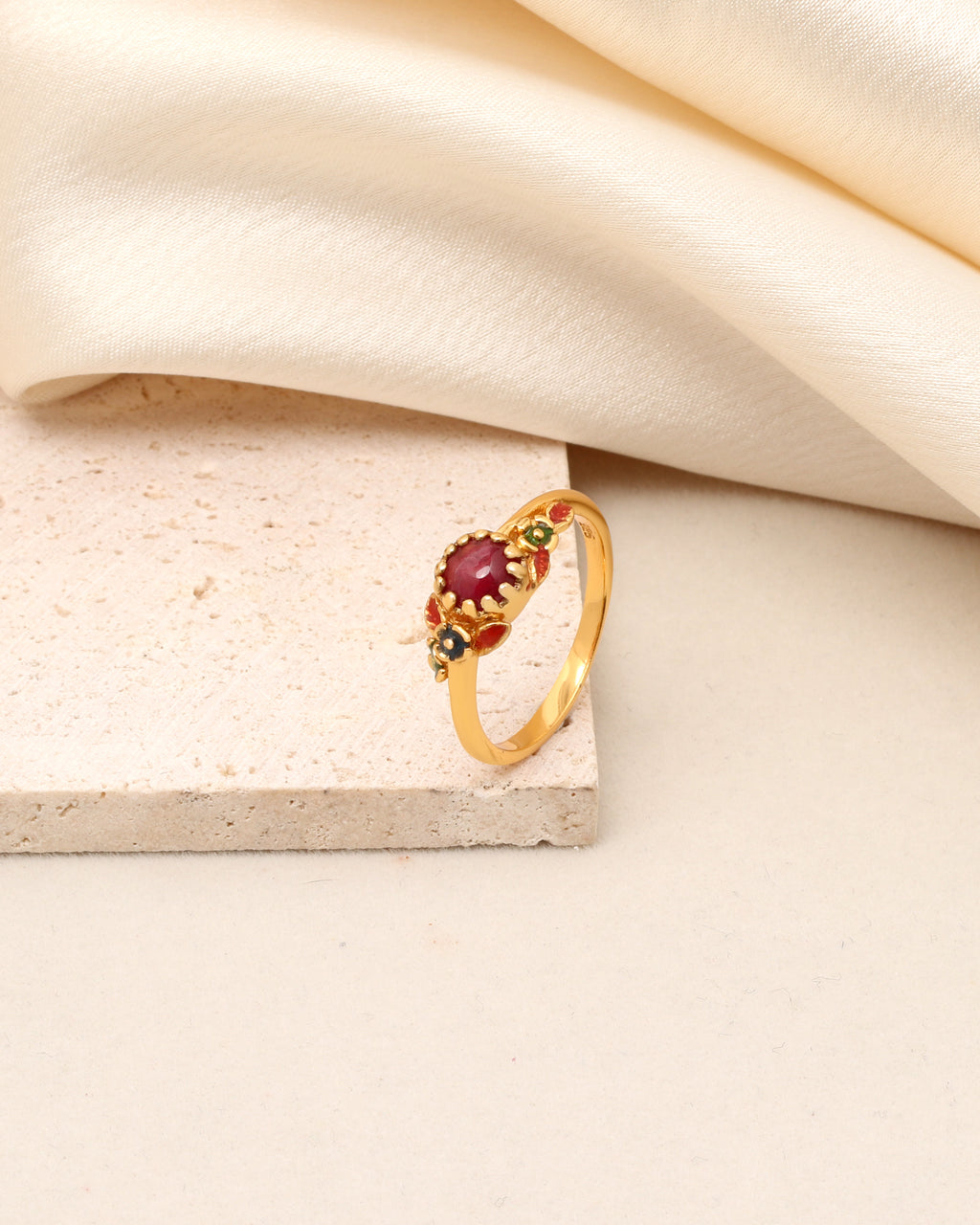 A gold ring with a central oval red gemstone and small green and red accent stones, displayed on a textured beige stone block with draped cream fabric in the background.