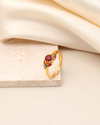 A gold ring with a central oval red gemstone and small green and red accent stones, displayed on a textured beige stone block with draped cream fabric in the background.