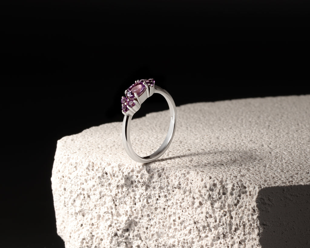 A silver ring with a marquise-cut pink gemstone in a prong setting, displayed on a textured white stone block against a solid black background.