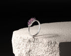 A silver ring with a marquise-cut pink gemstone in a prong setting, displayed on a textured white stone block against a solid black background.