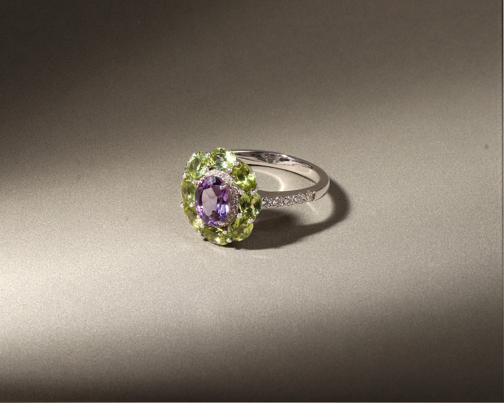 A silver ring featuring a central oval purple amethyst gemstone surrounded by a halo of light green peridot gemstones, with a small row of clear accent stones on the band, set against a dark gray background.