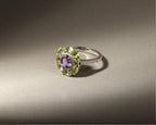A silver ring featuring a central oval purple amethyst gemstone surrounded by a halo of light green peridot gemstones, with a small row of clear accent stones on the band, set against a dark gray background.