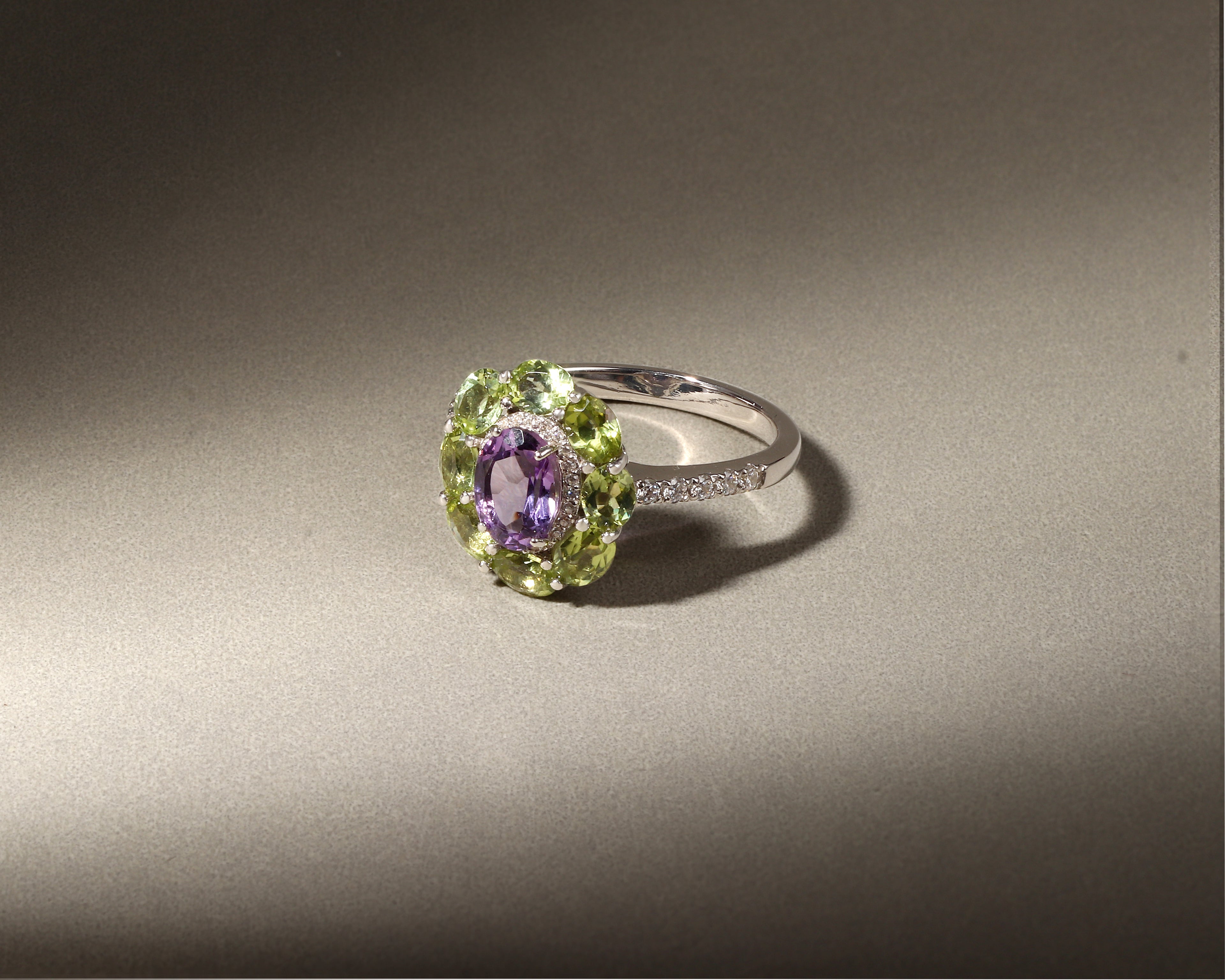 A silver ring featuring a central oval purple amethyst gemstone surrounded by a halo of light green peridot gemstones, with a small row of clear accent stones on the band, set against a dark gray background.