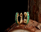 A pair of gold huggie hoop earrings adorned with round green emerald-cut gemstones, displayed on a textured piece of wood against a solid black background.