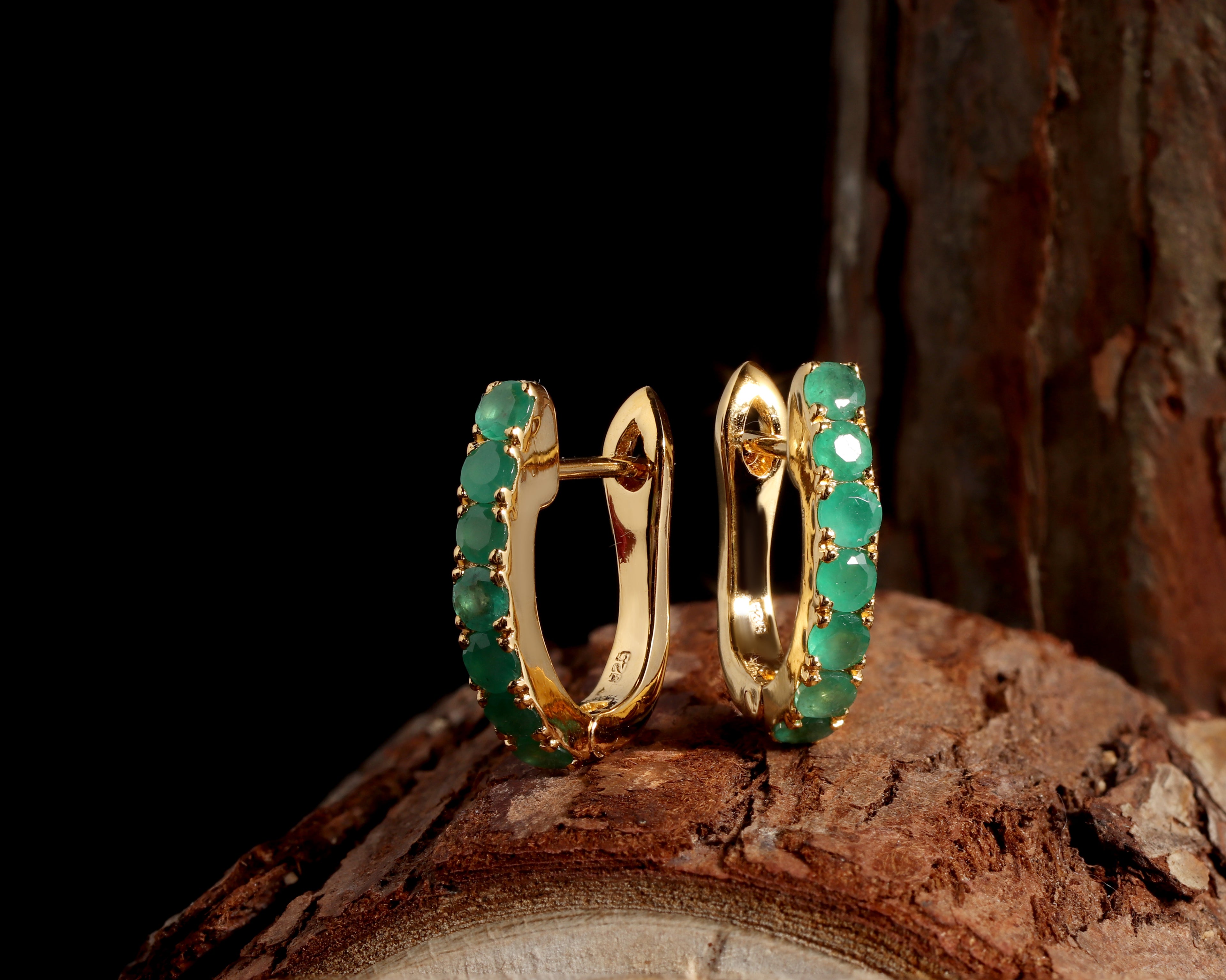A pair of gold huggie hoop earrings adorned with round green emerald-cut gemstones, displayed on a textured piece of wood against a solid black background.