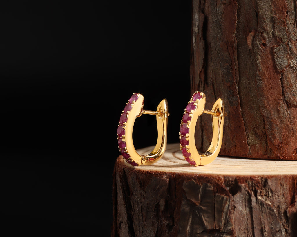 A pair of gold huggie hoop earrings with a row of small pinkish-purple stones along the front, displayed on a wooden log against a black background.