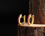 A pair of gold huggie hoop earrings with a row of small pinkish-purple stones along the front, displayed on a wooden log against a black background.
