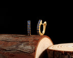 A pair of gold hoop earrings with dark blue gemstones, displayed on a rustic wood log against a solid black background.