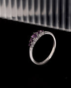 A silver band ring featuring a curved row of small round purple gemstones, displayed against a black background with a blurred ribbed white surface in the upper background.