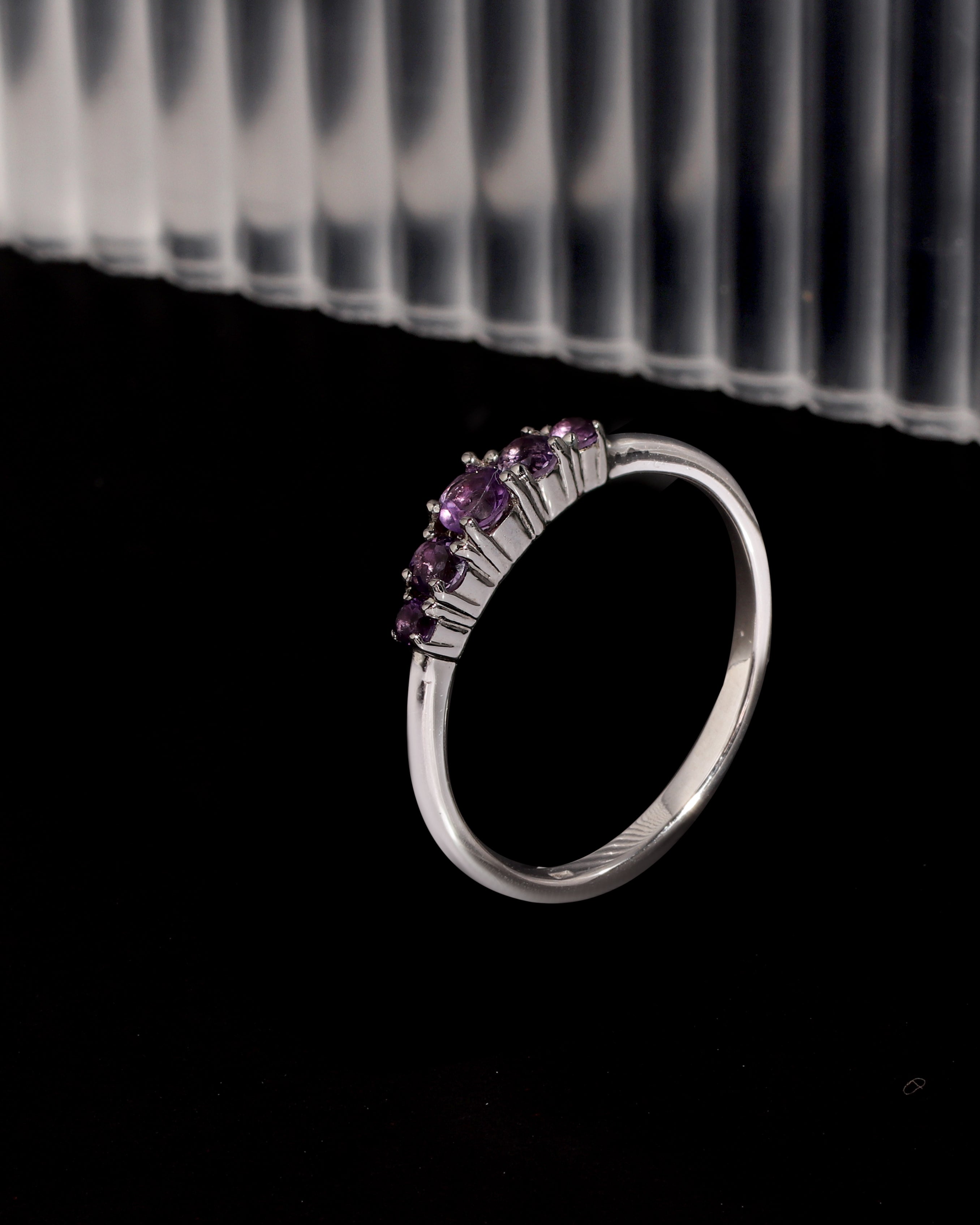 A silver band ring featuring a curved row of small round purple gemstones, displayed against a black background with a blurred ribbed white surface in the upper background.