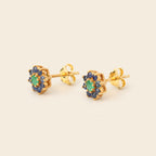 A pair of gold stud earrings featuring a central green stone surrounded by a halo of small blue stones, shown on a plain beige background.