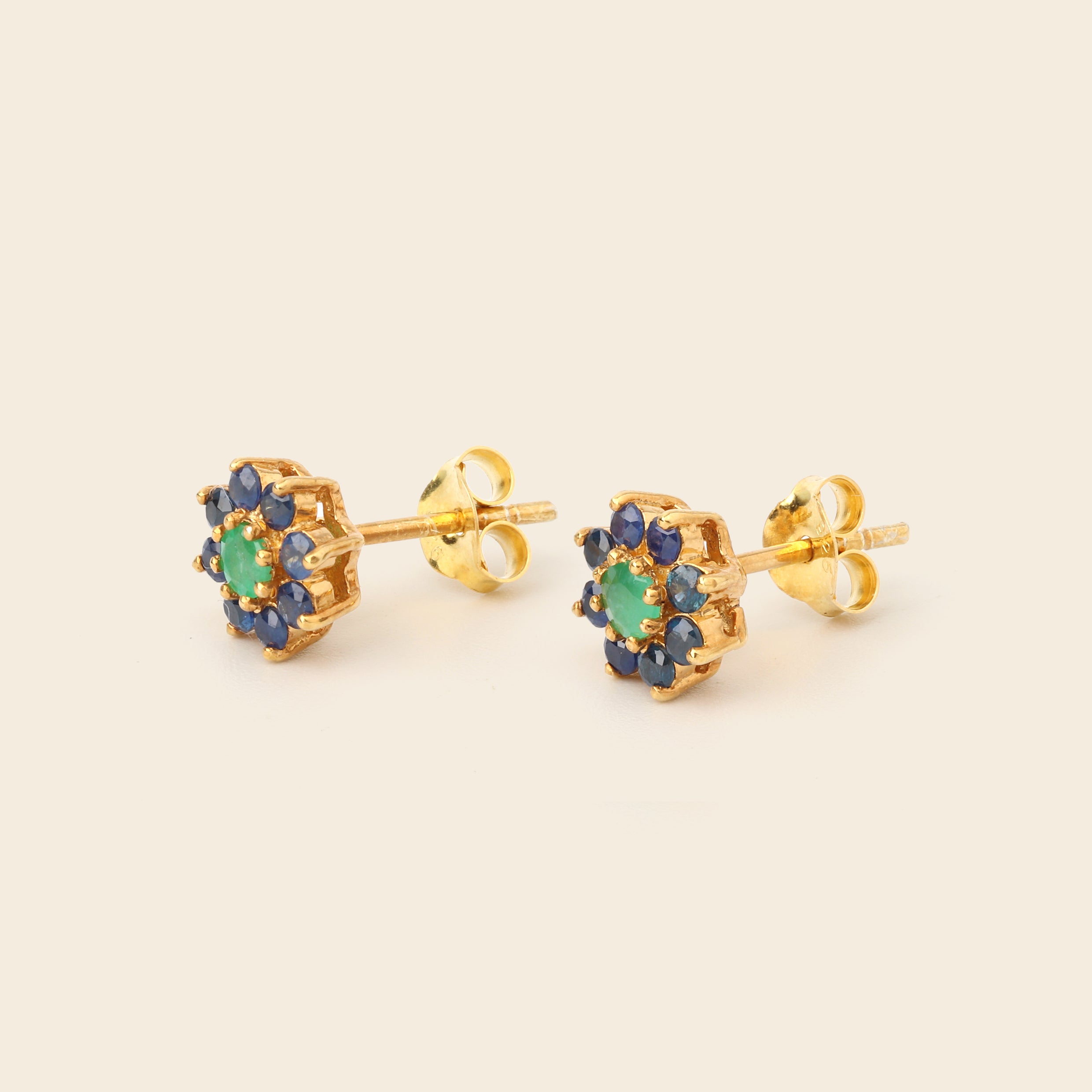 A pair of gold stud earrings featuring a central green stone surrounded by a halo of small blue stones, shown on a plain beige background.