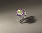 A silver ring featuring a central oval green gemstone surrounded by a halo of round purple gemstones, with small clear stones along the band, set against a neutral gray background.