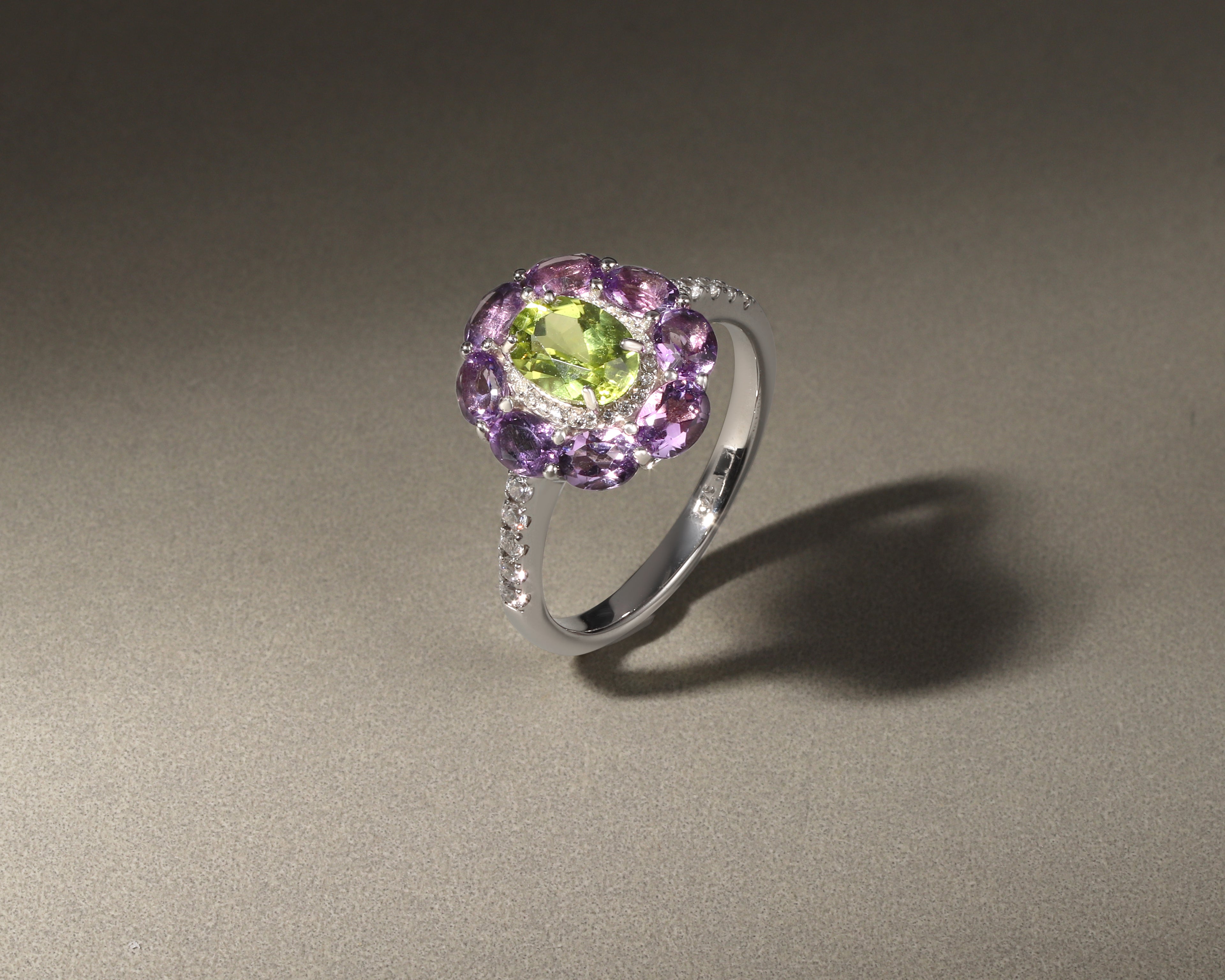 A silver ring featuring a central oval green gemstone surrounded by a halo of round purple gemstones, with small clear stones along the band, set against a neutral gray background.