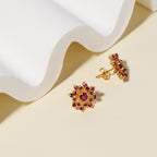 A pair of gold flower-shaped stud earrings with red gemstone accents, displayed on a beige surface with a white wavy prop.