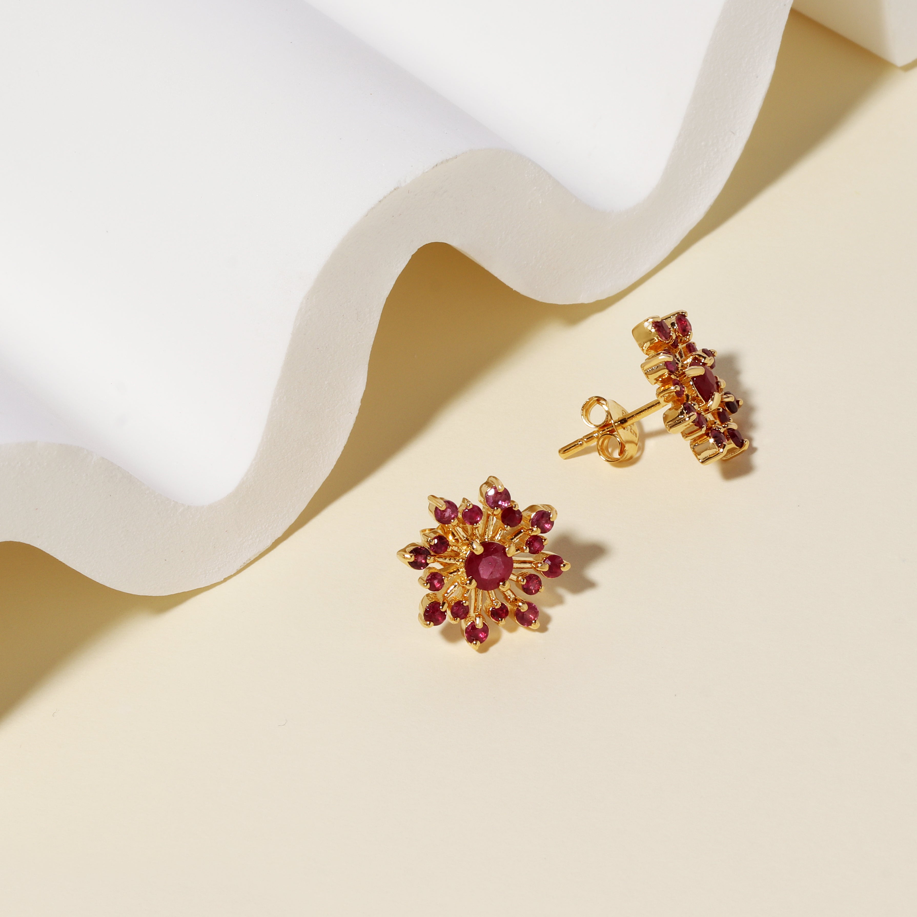 A pair of gold flower-shaped stud earrings with red gemstone accents, displayed on a beige surface with a white wavy prop.