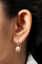 A close-up of a person's ear wearing a delicate gold huggie hoop earring with a small rose-shaped dangling charm.