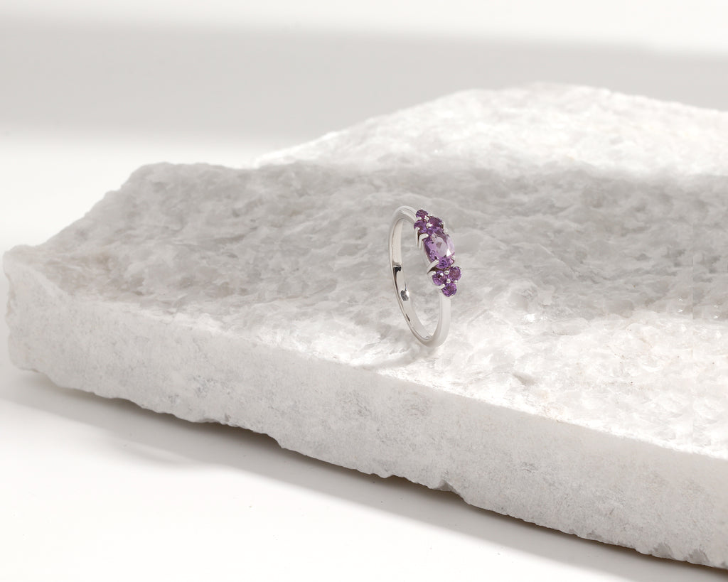 A silver band ring with a small cluster of purple gemstones, displayed on a textured white stone slab against a clean white background.