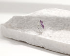 A silver band ring with a small cluster of purple gemstones, displayed on a textured white stone slab against a clean white background.