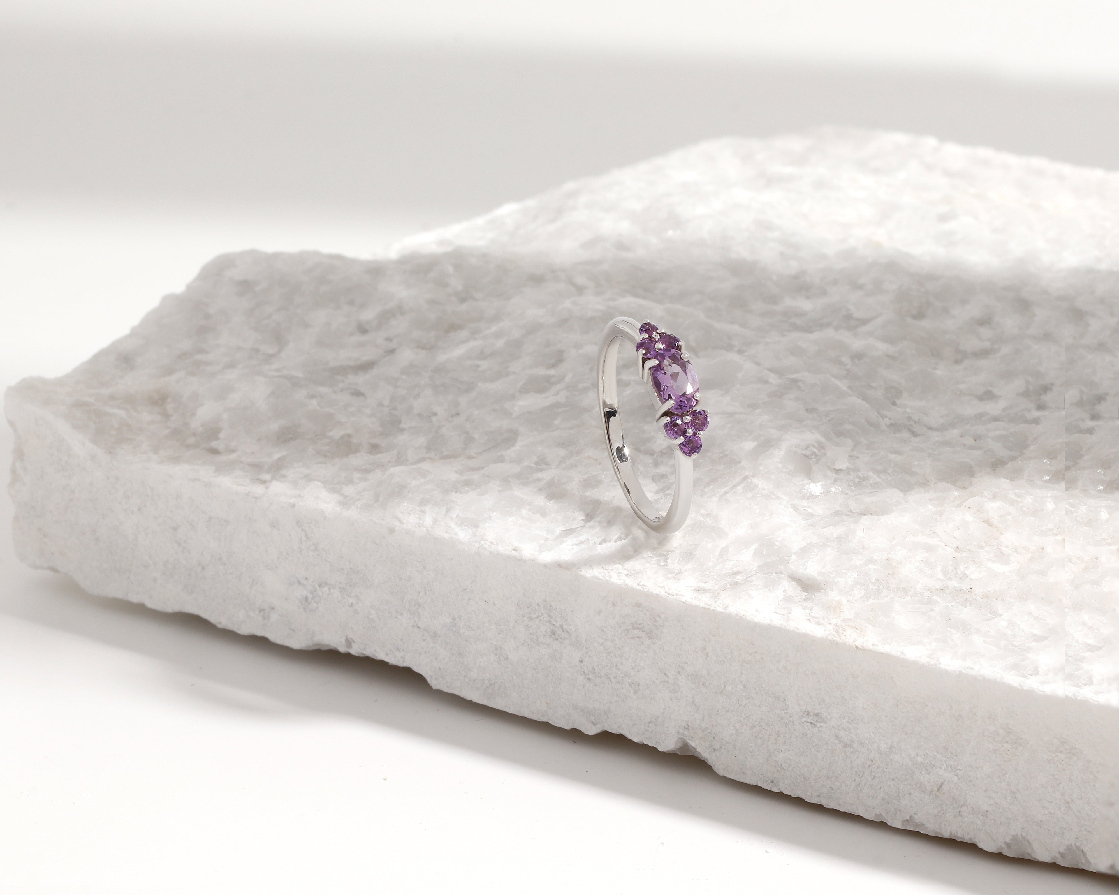 A silver band ring with a small cluster of purple gemstones, displayed on a textured white stone slab against a clean white background.