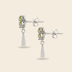 A pair of silver drop earrings featuring a small cluster of light green gemstones in a textured bezel setting, with a teardrop-shaped clear stone dangling below, shown on a light beige background.