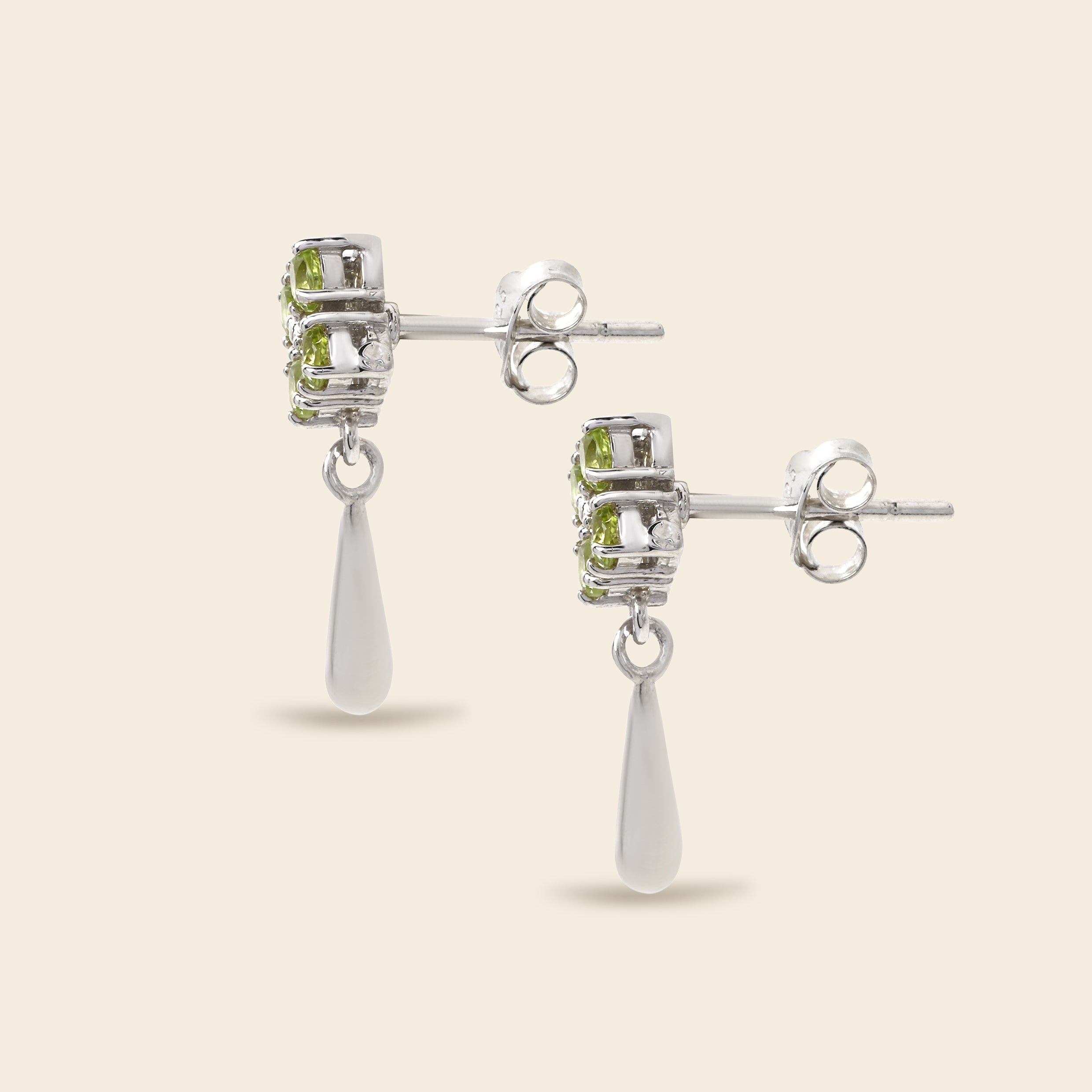 A pair of silver drop earrings featuring a small cluster of light green gemstones in a textured bezel setting, with a teardrop-shaped clear stone dangling below, shown on a light beige background.