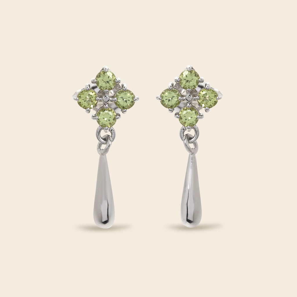 A pair of silver dangle earrings featuring a four-stone floral cluster in light green peridot gemstones, with a teardrop-shaped silver drop below, set against a solid beige background.