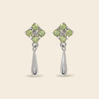 A pair of silver dangle earrings featuring a four-stone floral cluster in light green peridot gemstones, with a teardrop-shaped silver drop below, set against a solid beige background.