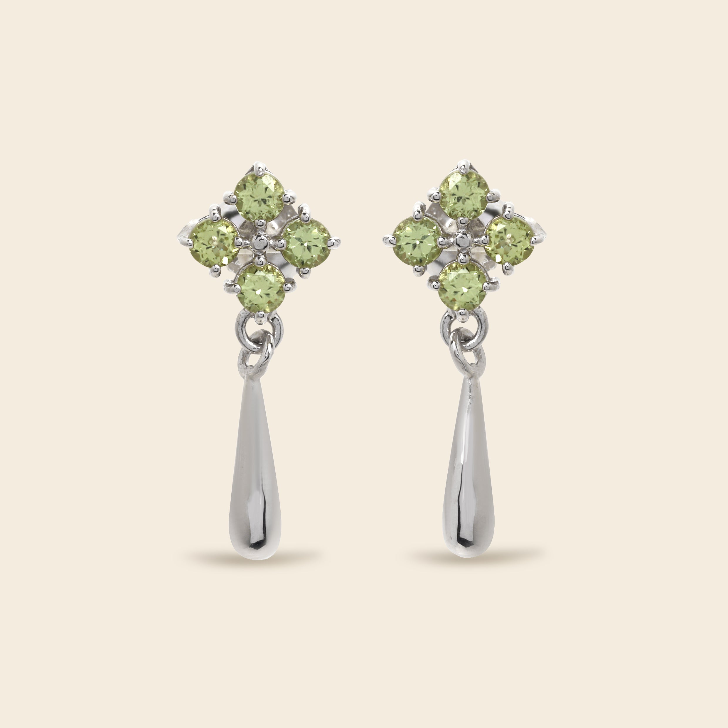 A pair of silver dangle earrings featuring a four-stone floral cluster in light green peridot gemstones, with a teardrop-shaped silver drop below, set against a solid beige background.