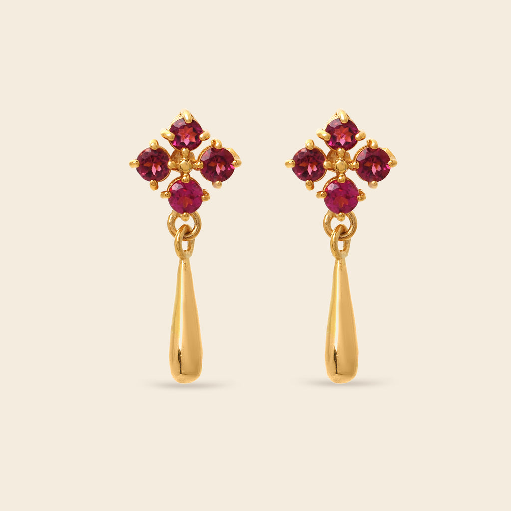 A pair of gold dangle earrings featuring a four-stone floral cluster at the top with deep red gemstones and a teardrop-shaped gold drop below, set against a solid light beige background.