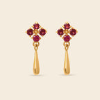 A pair of gold dangle earrings featuring a four-stone floral cluster at the top with deep red gemstones and a teardrop-shaped gold drop below, set against a solid light beige background.