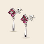 A pair of silver dangle earrings featuring a cluster of five round-cut pink gemstones in a floral setting, with a polished silver teardrop drop hanging below, shown against a solid light beige background.