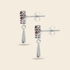 A pair of silver drop earrings with a rectangular cluster of small red gemstones and a teardrop-shaped dangling drop, shown on a light beige background.