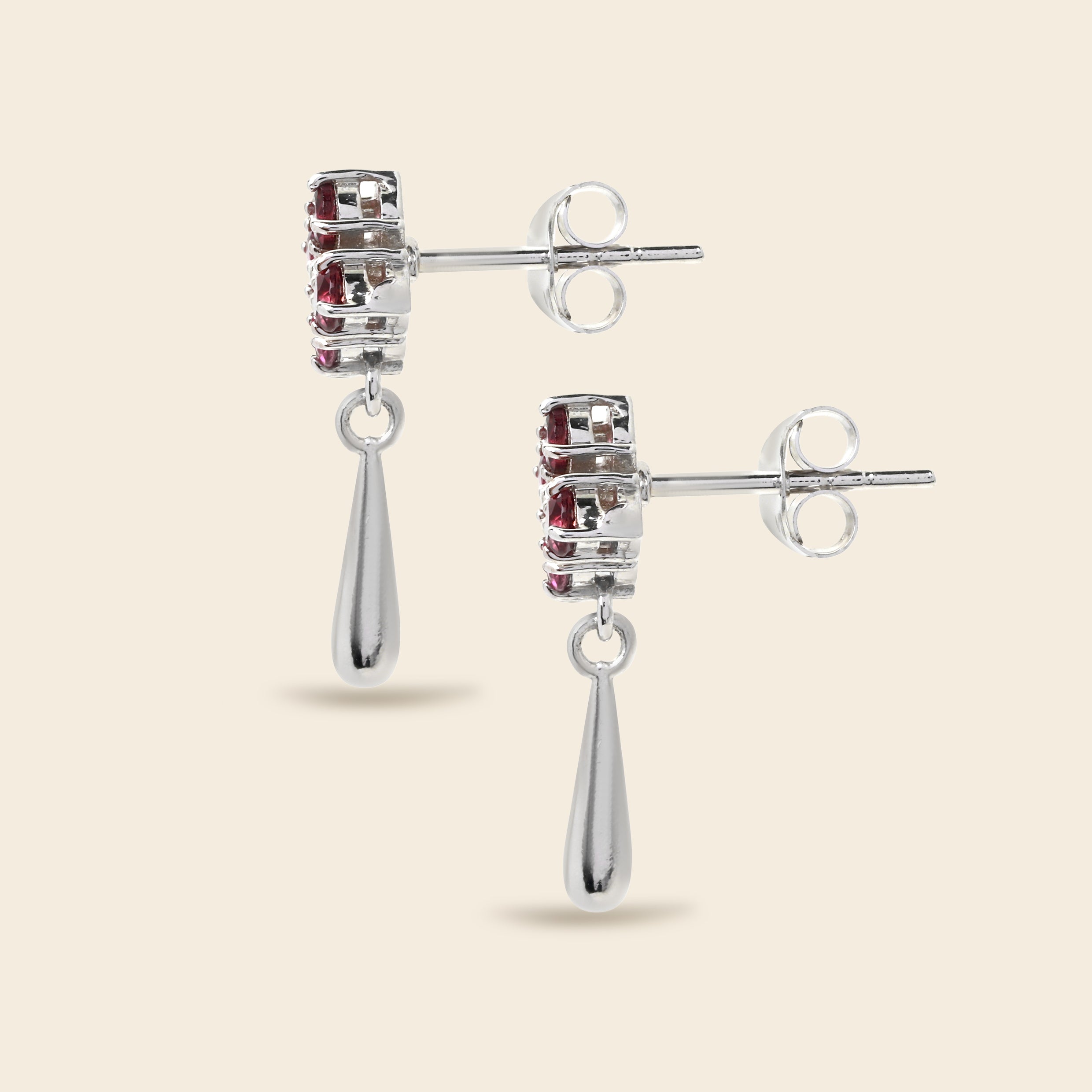 A pair of silver drop earrings with a rectangular cluster of small red gemstones and a teardrop-shaped dangling drop, shown on a light beige background.
