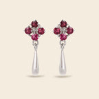 A pair of silver dangle earrings featuring a four-stone floral cluster at the top with four round pinkish-red gemstones and a teardrop-shaped silver drop below, set against a solid light beige background.