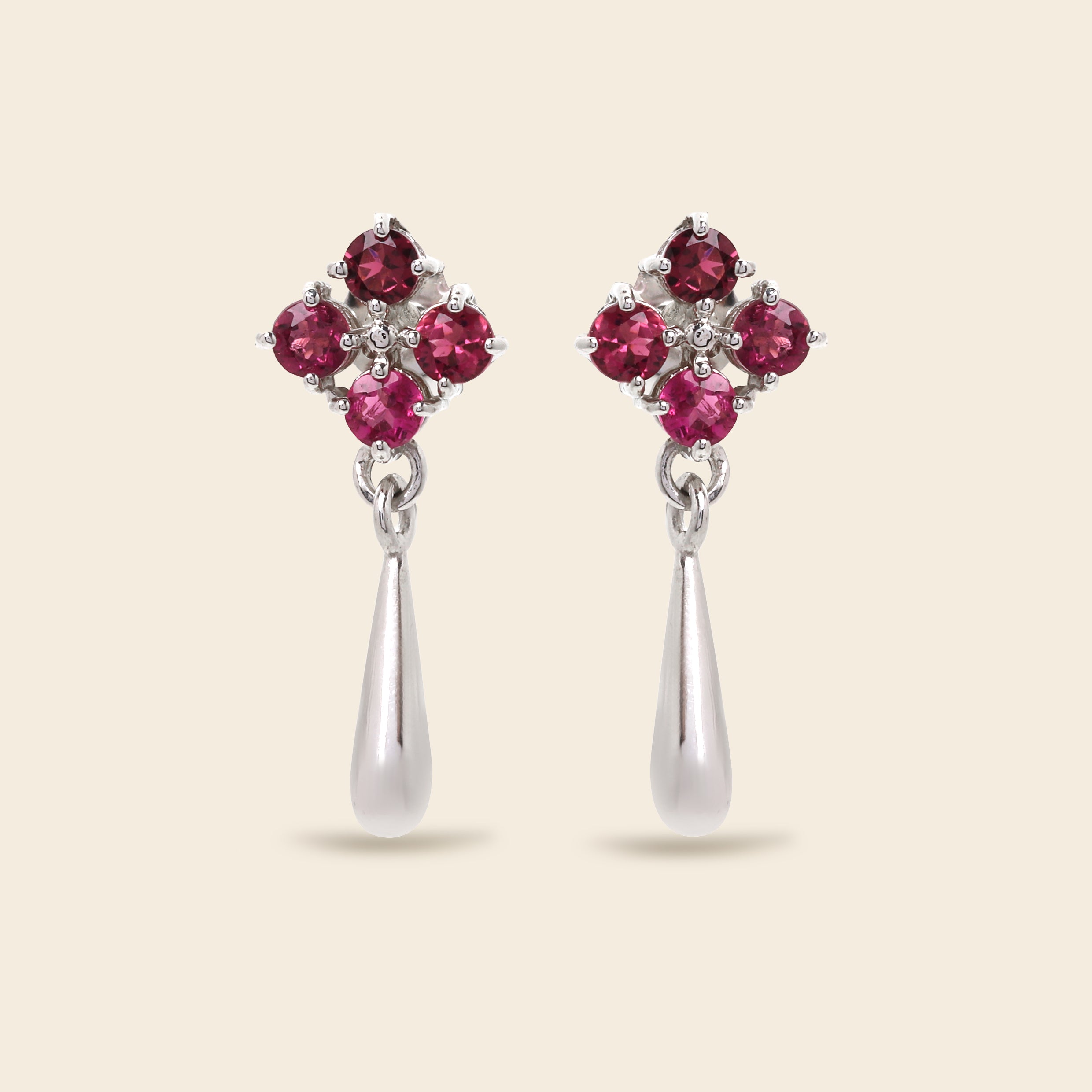 A pair of silver dangle earrings featuring a four-stone floral cluster at the top with four round pinkish-red gemstones and a teardrop-shaped silver drop below, set against a solid light beige background.