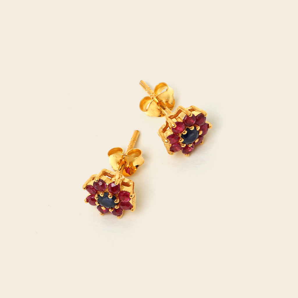 A pair of gold stud earrings with dark red gemstone clusters and a central dark blue stone, displayed on a light cream background.