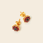 A pair of gold stud earrings with dark red gemstone clusters and a central dark blue stone, displayed on a light cream background.