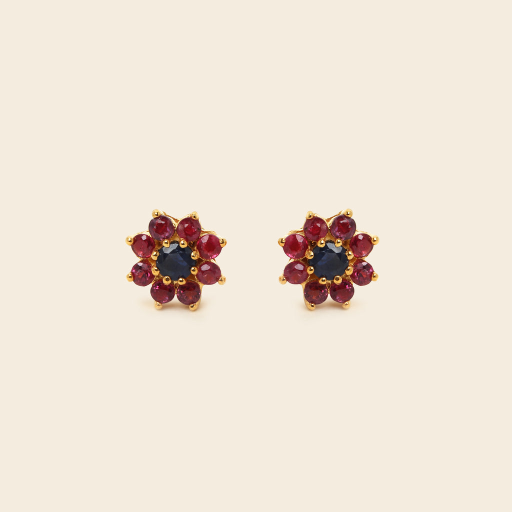 A pair of gold flower-shaped stud earrings featuring a dark blue center stone surrounded by red gemstones, set against a solid cream background.